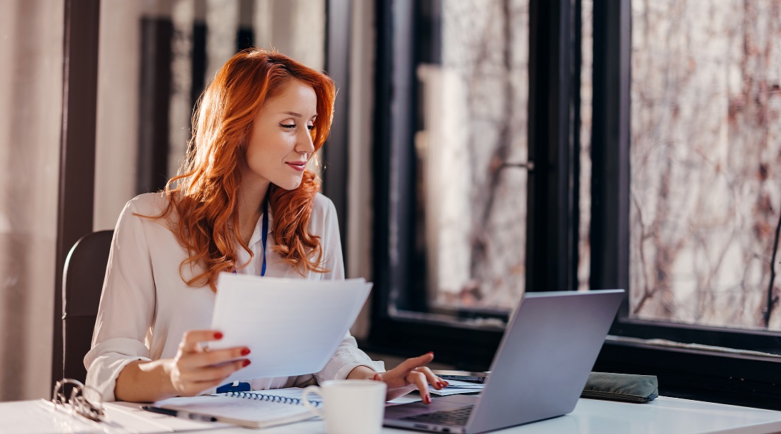 happy smiling businesswoman in formal wear sitting at desk in modern office in front of laptop and doing paperwork